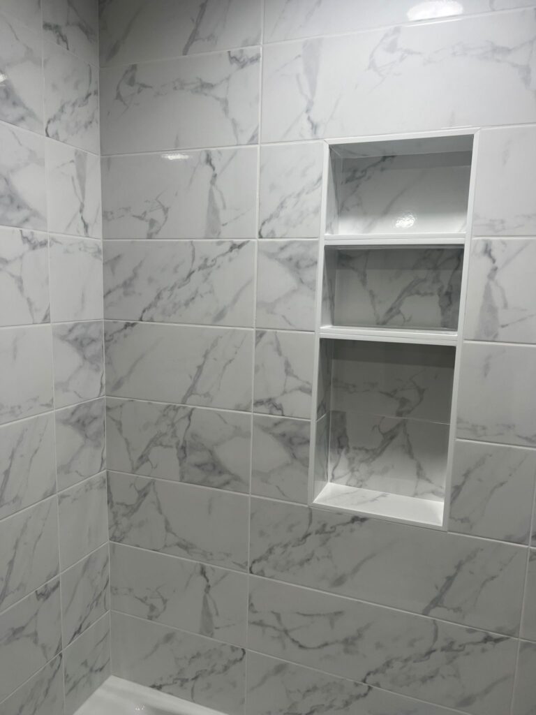 A bathroom with a white bathtub and shower head, surrounded by white tiles with a gray marble pattern.
